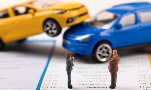 Two miniature people standing on an accident report form and looking at two toy cars positioned as if they collided.