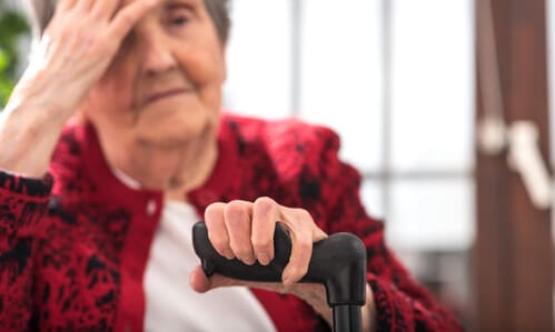 A partially out-of-focus shot of an elderly woman holding a cane in her left hand, and her head in her right hand.