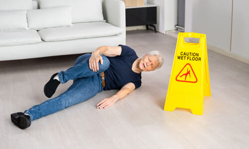 An old man lying on his side and holding his knee next to a wet floor sign after a slip-and-fall accident.