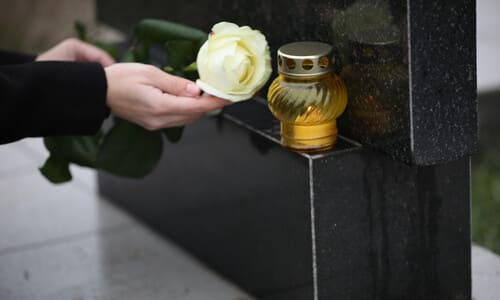 A mourning woman's hands in black sleeves offering a white rose to a black tombstone with a lit candle.