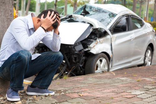 A driver crouches with his head in hand after a drunk driving accident. Silver car with front end damage pictured in background.