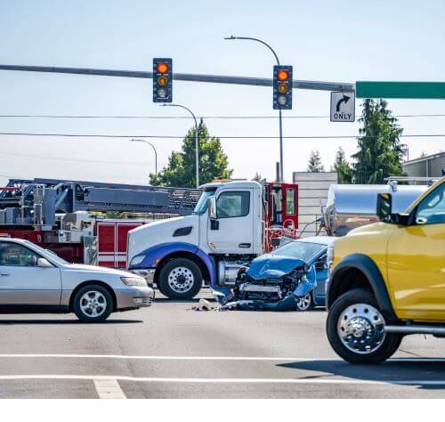 A blue car with extensive front-end damage after an accident with a truck. Emergency personnel pictured in the accident intersection.