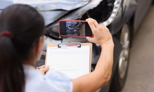 An over-the-shoulder view of a female insurance adjuster recording footage of a damaged car on her phone.
