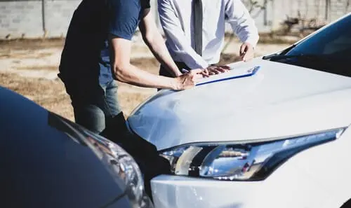 Two men assessing the collision damage of two cars and filling out a form on a clipboard on the hood of one of them.