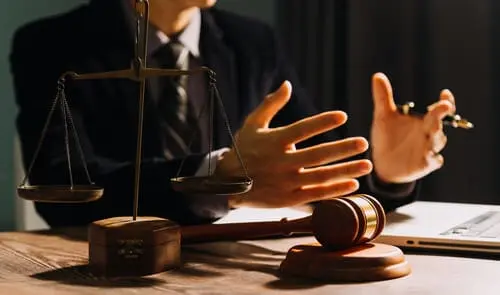 A lawyer behind a desk with a laptop, gavel, and scales, and explaining something to an unseen client.