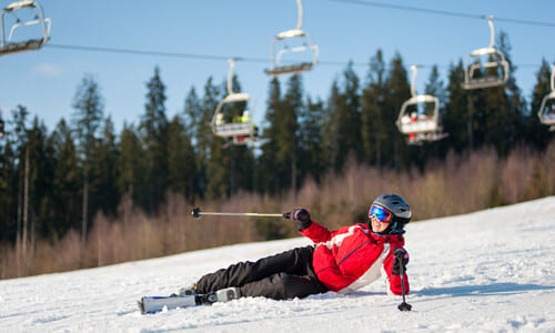 A woman in red outerwear falling over on her side while skiing down a slope.