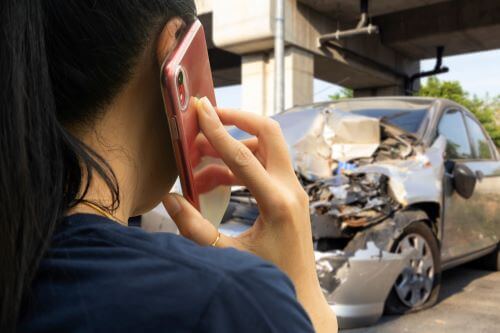 A woman is on the phone with a damaged vehicle in the background after a car accident.