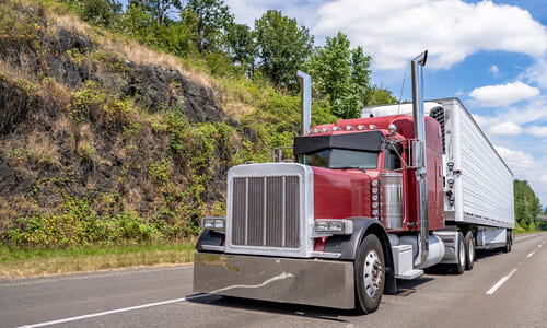 A cherry-red semi truck driving along a mountain road on a sunny day.
