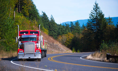A red big rig driving on a forested mountain road pulling an end dump trailer.