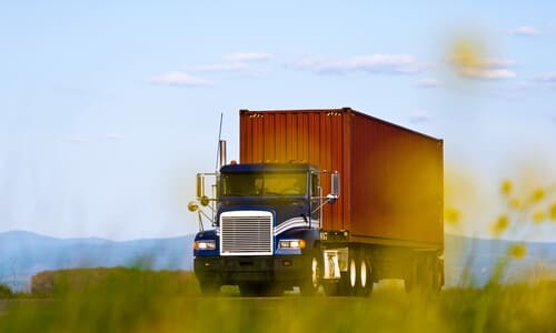 A dark blue semi truck pulling a red trailer filled with cargo on a country road.