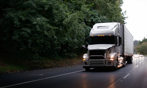 A large white delivery truck with headlights on driving on a rainy forested mountain road in the afternoon.