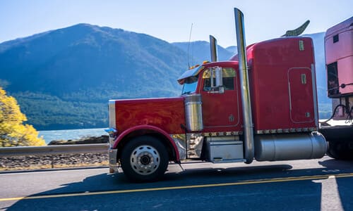 A red semi truck pulling its matching trailer behind it on a road with a mountain lake in the background.