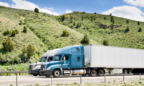 A blue semi truck pulling a grey trailer along a road next to a hillside.