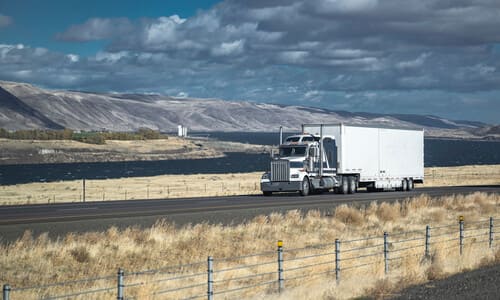 A white semi truck and its trailer driving along an open road with a wide river in the background.