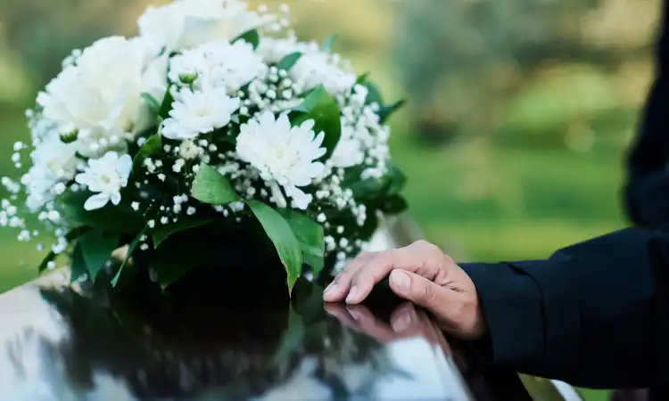 A mourner at a funeral with a hand on a casket placed next to a bouquet of white flowers.