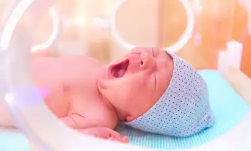 A newborn baby boy yawning inside an incubator in a hospital nursery.