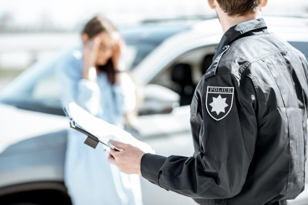 Policeman issuing a fine for violating the traffic rules to a young upset woman driver stadning near the car on the background