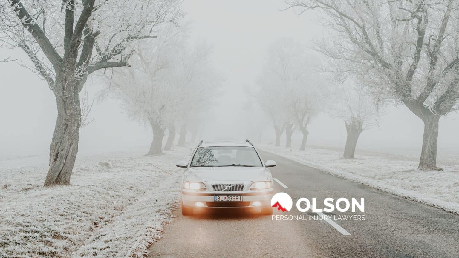 a light-colored car driving on a tree-lined, snowy road.