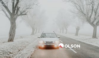 a light-colored car driving on a tree-lined, snowy road.