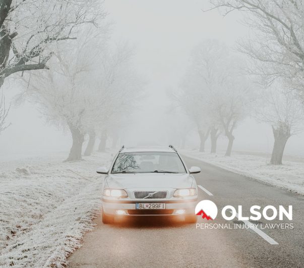 a light-colored car driving on a tree-lined, snowy road.