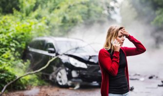 A frustrated young woman with smartphone by the damaged car after a car accident, making a phone call.