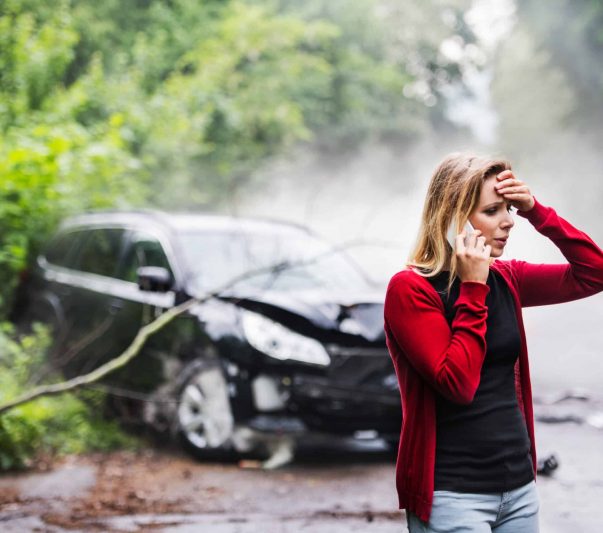 A frustrated young woman with smartphone by the damaged car after a car accident, making a phone call.