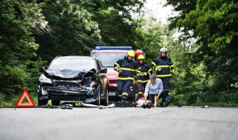 Firefighters running to rescue a woman lying unconscious on the road after a car accident. A mature man holding her hand, checking her pulse. Copy space.